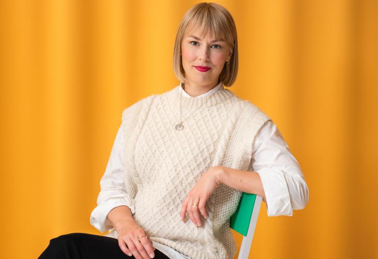 Kuvassa Ida Kukkapuro istumassa tuolilla puolivartalokuvassa. Hänellä on valkoinen pusero päällään. Hän nojaa vasemmalla kädellään tuolin käsinojaan.  Taustalla on keltainen verho.