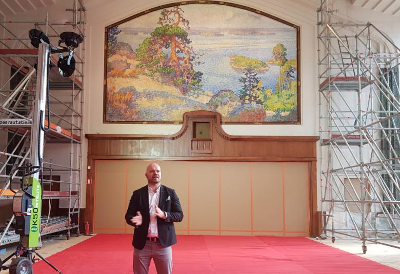 Kuvassa on taustalla konservoitu Järnefeltin Maisema Kolilta päärautatieaseman ravintolasalissa. Teoksessa on kalliota ja mäntyjä sekä oikealla järveä. Salissa on vaalea takaseinä, jonka yläosassa on teos. Molempien seinien vierellä on rakennustelineitä.