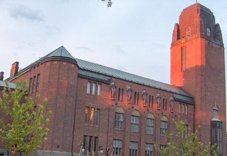 Kuvassa on Joensuun kaupungintalo, joka on punatiilinen ja siinä on kolme kerrosta. Ikkunat ovat eri muotoisia ja mm. kapeita pystysuoria. Oikealla on torni.