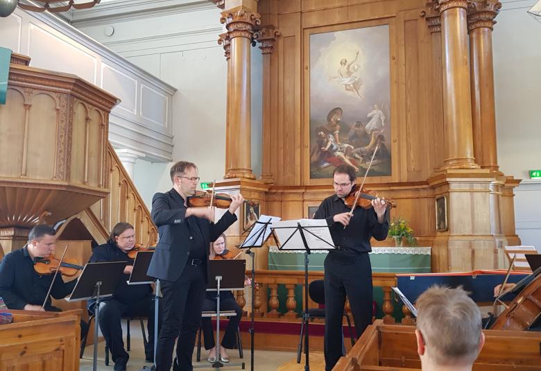 Kuvassa ovat soittamassa Kaakon Kamarimusiikin soittajat edessä soittamassa viulua vasemmalla Jukka Merjanen ja oikealla Petteri Iivonen. Taustalla on vaalean ruskeat pylväikot ja alttaritaulu sekä oikealla saarnastuoli.