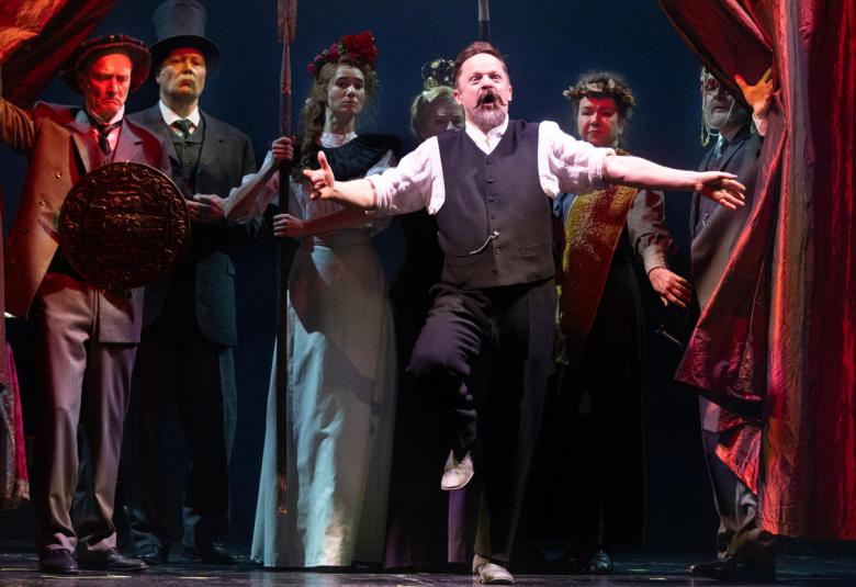 Kuvassa on näyttämöllä kuusi näyttelijää ja miehet ovat puvuissa ja naisella on pitkä hame päällään. Keskellä on seitsemäs näyttelijä, mies on mustassa liivssä ja housuissa sekä valkeassa paidassa. Hän pitä käsiään vaakatasossa ja oikeaa polvea ylhäällä. Reunimmaiset henkilöt pitävät punaisia samettiverhoja syrjässä.