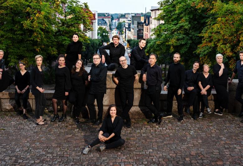 Kuvassa on Key Ensemblen kuorolaisia levittäytyneenä ja seisomassa sekä osittain istumassa takanaan vihreitä lehtipuita sekä kiviaitaa. 