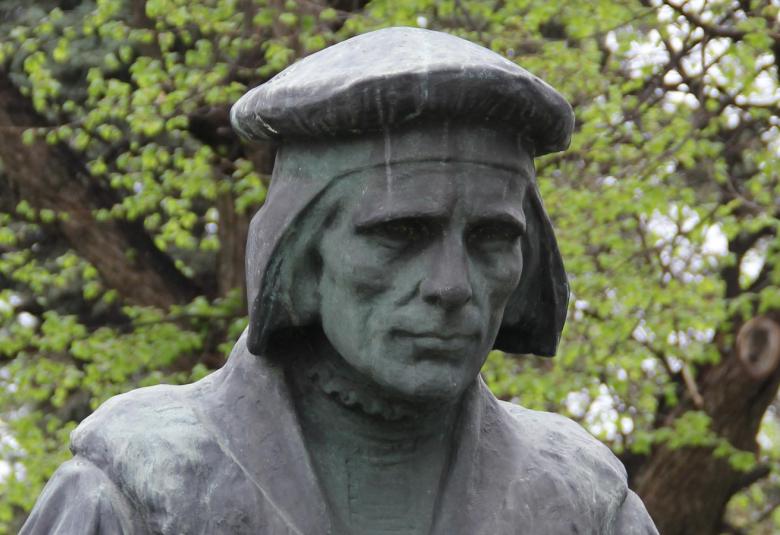 Kuvassa on Mikael Agricolan patsaasta Agricolan pää. Se on hieman kumartuneena alaspäin. Patsas on mustaa kiveä ja taustalla on vihreää lehtipuun lehvästöä.