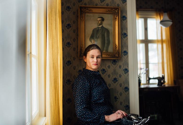 Kuvassa on vasemmalla iso ikkuna ja keltainen verho ja sen vieressä seisoo Paula Nivukoski mustassa mekossa. Taustalta näkyy vanhanaikaista kalustusta ja huonetilaa.