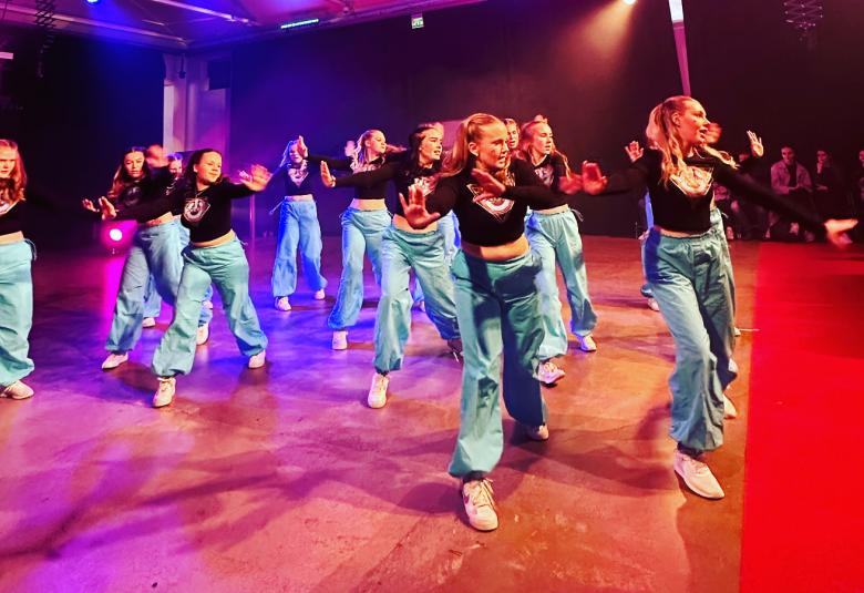 Kuvassa on nuoria tanssijoita lavalla, joka on punasävyinen. Tanssijoilla on vaaleansiniset housut ja mustat t-paidat. 