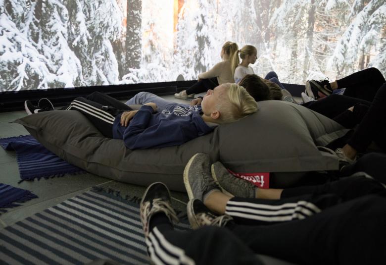 Kuvassa on tila, jossa on nuoria hämärässä löhöpatjojen päällä katsomassa seinällä olevaa isoa taideteosta, jossa on lumisia kuusenoksia ja metsää.