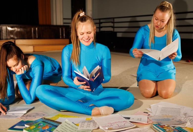 Kuvassa on kolme tyttöä vaalean sinisissä jumppa-asuissa.He ovat lattialla istumassa ja tutkimassa lehtiä sekä kirjoja. Lattialla on molempia ja taustalla näkyy jumppasalia.