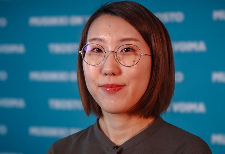 Kuvassa on Mioko Yokoyama kasvokuvassa. Hänellä on pyöreät silmälasit ja ruskea suora tukka. Taustalla on sinistä seinää ja siinä on valkoista himmeää tekstiä.