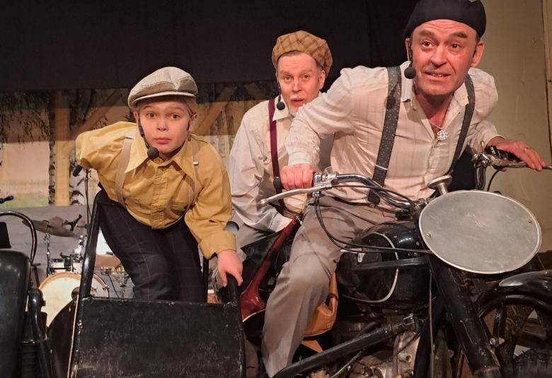 Kuvassa on mies vanhan moottoripyörän selässä ja hän on kumartuneena eteenpäin. Hänen takanaan kanssaan on kaksi poikaa lippalakessa moottoripyörän takapenkillä. 