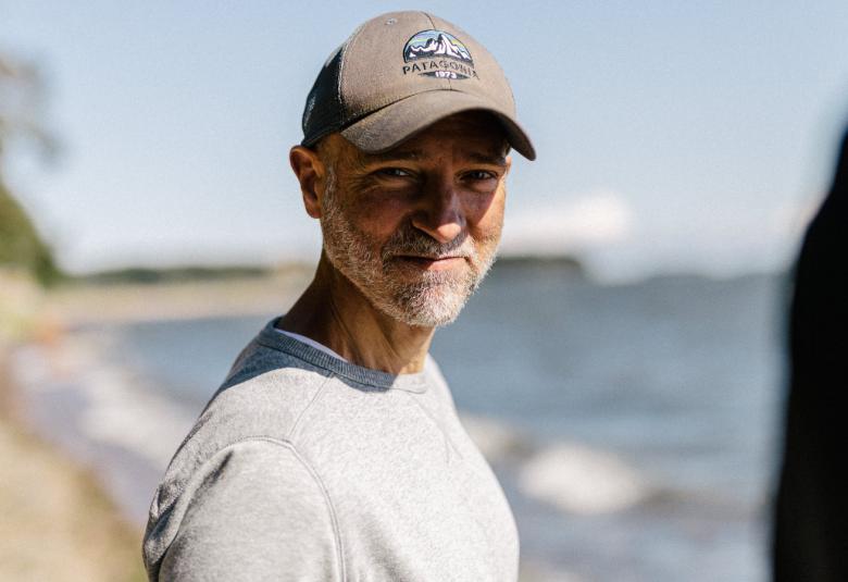 Kuvassa on Aku Louhimies puolivartalokuvassa ja hänellä on lippalakki päässään ja siitä tulee puolivarjo kasvoille. Taustalla näkyy järveä ja rantaa kesällä.