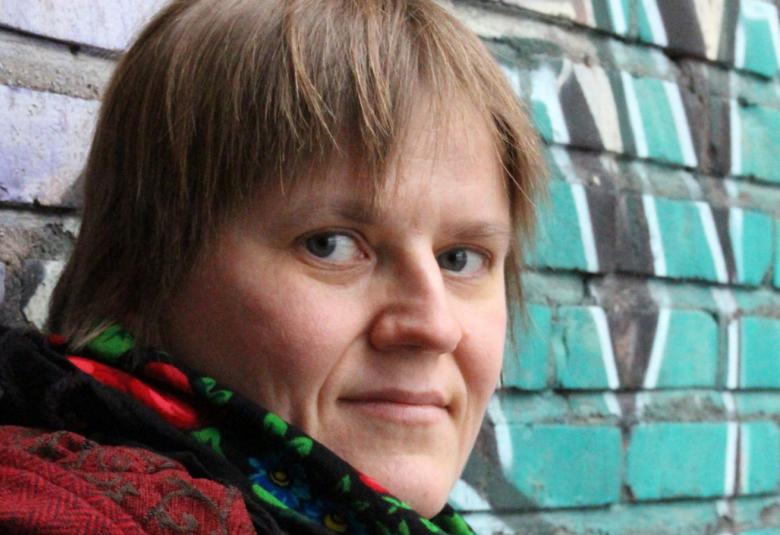 Kuvassa on Anne Leinonen kasvokuvassa ja hänellä on kukallinen huivi kaulassaan. Oikealla näkyy betoniseinän vihreä graffiti. 