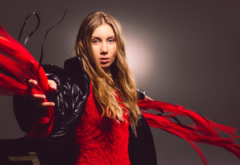 Kuvassa on Ella Marie vauhdikkaassa asennossa ja hänellä on käsissään punaiset huivit sekä huivi kaulassaan. Hänellä on pitkät hiukset. Tausta on tumma.