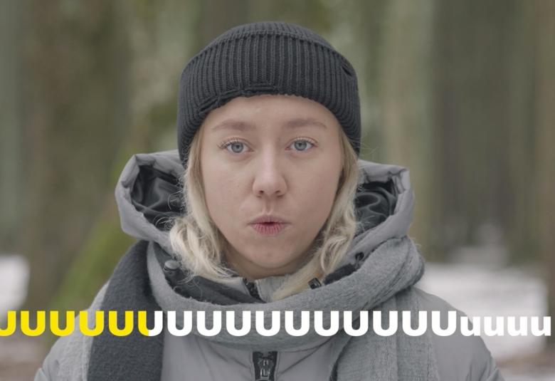 Kuvassa on nuori nainen puolivartalokuvassa ja hänellä on harmaa huppari sekä musta pipo. Hänellä on suu u-asennossa.  Alaosassa on kirjaimen u jono ja ensimmäiset kuusi ovat keltaisia ja loput valkoisia.