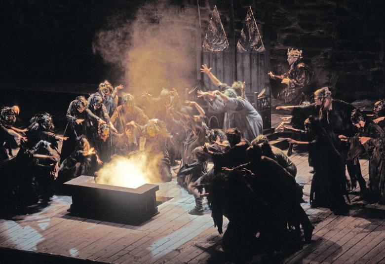Kuvassa on tummalla näyttämöllä tummissa kaavuissa tulen ympärille kumartuneet hahmot, jotka osoittavat tulta kohti kädellään.