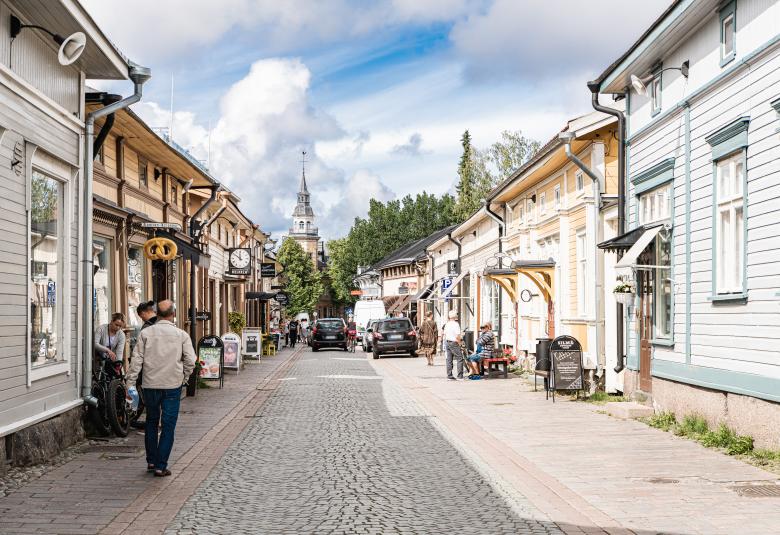 Kuvassa on katunäkymä Vanhalta Raumalta ja siin on molemmin puolin katua puutaloja. Keskellä on mukulakivikatu ja siinä kävelee ihmisiä. Talot ovat vaaleita. Kuva on otettu kesällä.