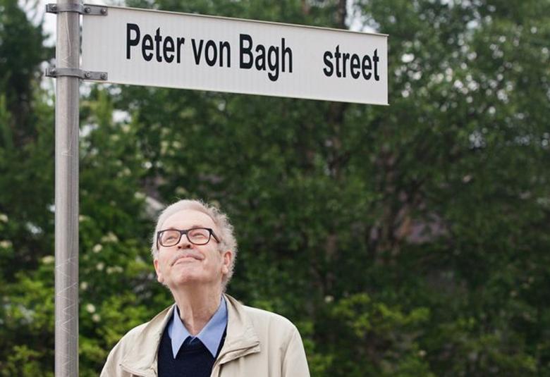 Kuvassa on Peter von Bagh alhaalla ja hän katsoo yläpuolellaan olevaa katukylttiä, jossa lukee Peter von Bagh Street. Taustalla on vihreää lehtipuumetsää.