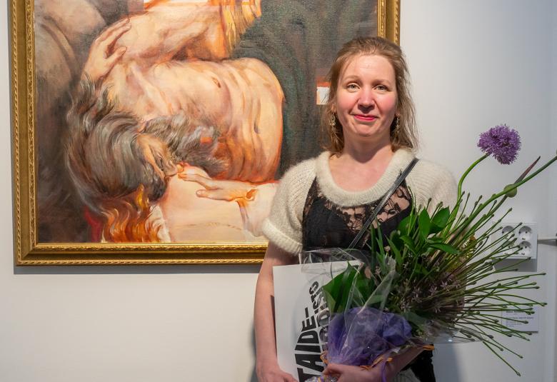 Kuvassa on Anna Tereshko seisomassa kukkakimppu kädessään ja hänellä on vaalea villapusero päällään.  Taustalla on isokokoinen taulu ja siitä näkyy osa, jossa on vaalean ruskeaa ja turkoosia.