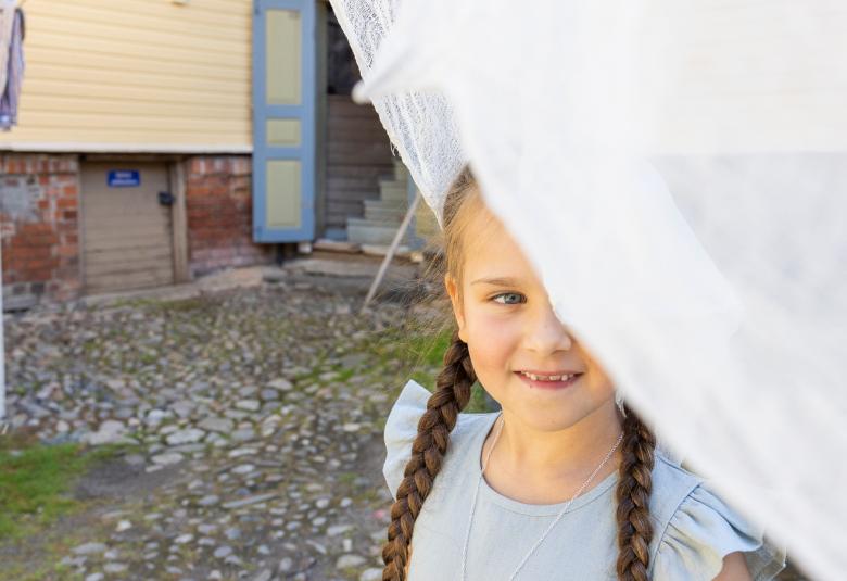 Kuvassa on oikealla vinosti valkoista verhoa ja sen alta näkyy lettipäisen pikkutytön kasvoja ja vaalen sinistä mekkoa. Taustalta näkyy kivistä pihaa ja keltaista seinää. 
