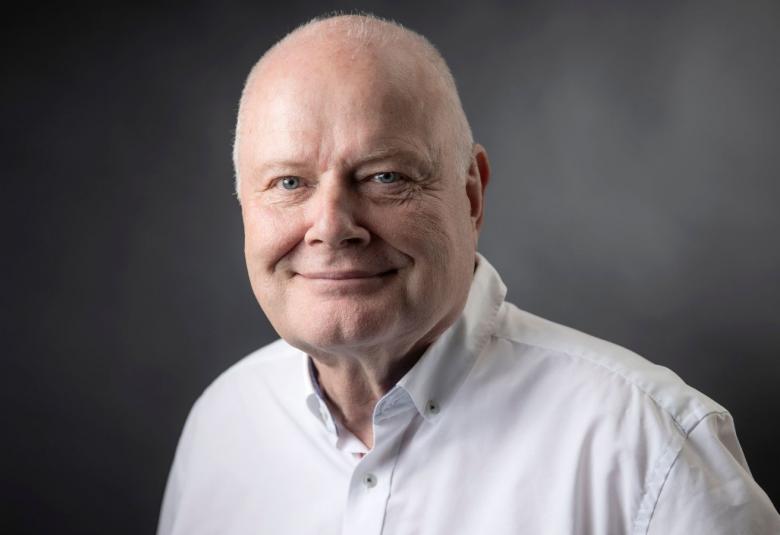 Kuvassa on Arvo Tuominen puolivartalokuvassa. Hänellä on valkoinen paita päällään ja ei ole hiuksia. Tausta on tumma.