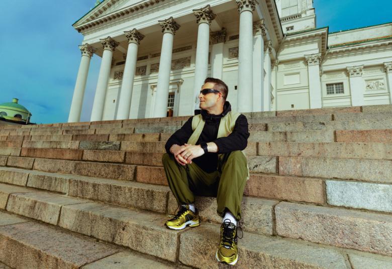 Kuvassa on Darude Helsingin tuomiokirkon portailla. Takana näkyy tuomiokirkon  valkoinenn fasadi. Darude istuu portailla rennosti vasen jalka edessä. Hänellä on mustat vaatteet päällään. Takana on sininen taivas.