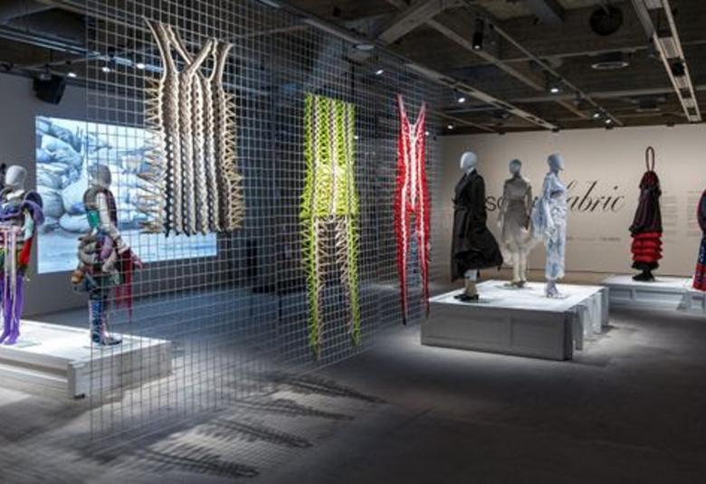 Kuvassa on näkymänäyttelysalista, jossa on vasemmalla kolme nallinukkea seisoassa rimpsullisissa vaatteissa. Keskellä on isosilmäistö verkkoa ja siinä roikkuu puvun mallisia mattoja. Oikealla on jalustalla lisää kolme mallinukkea ja kaksi pukua. 