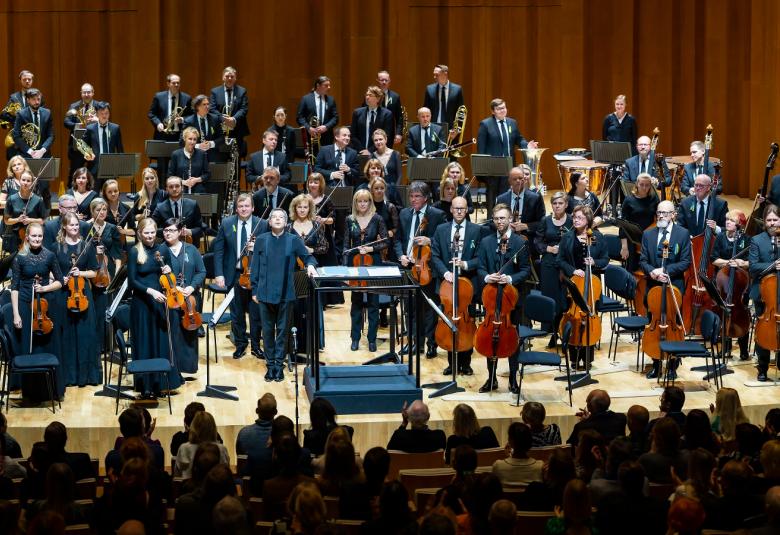 Kuvassa on sinfoniaorkesteri ERSO yhtieskuvassa ja he ovat seisomassa soittimineen lavalla. Heillä on tumman siniset puvut. Taustalla on ruskeaa verhoa.