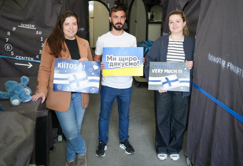 Kuvassa on vasemmalla ruskeassa jakussa nainen, jolla on kädessään kyltti, jossa lukee kiitos ja jossa on Suomen lippu ja lipun värejä. Keskellä on mies valkoisessa t-paidassa ja hänellä on kyltti, jossa on tekstiä ukrainaksi. Oikealla on nainen sinisessä jakussa ja valko-siniraitaisessa t-paidassa. Hänellä on kädessään kyltti, jossa lukee Thank you very much ja siinä on Suomen lippu ja Suomen lipun värit. Taustalla on varastohuonetta ja erilaisia pakattuja tavaroita.