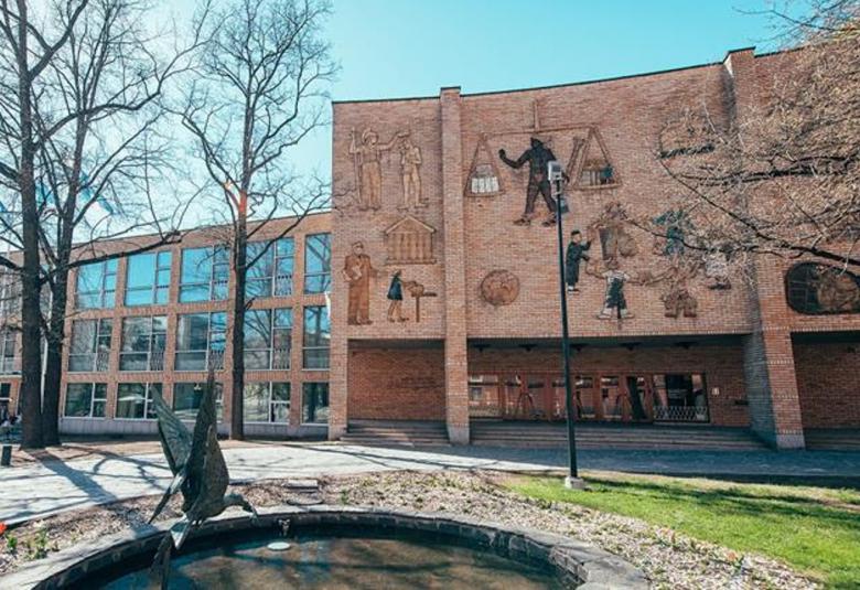 Kuvassa on Helsingin kauppakorkeakoulun entinen päärakennus, jossa on vasemmassa osassa isoja osittain ruudutettuja ikkunoita kolmessa rivissä ja oikealla puolella sisäänkäynnin julkisivu, jossa kohollaan olevia pintaveistoksia ja niissä ihmisiä työskentelemässä sekä esilaisia hahmoja.Rakennus on vaalean ruskeista tiilistä. Edessä on koristeallas, jossa on ylöspäin kurkottava vesilintuveistos.