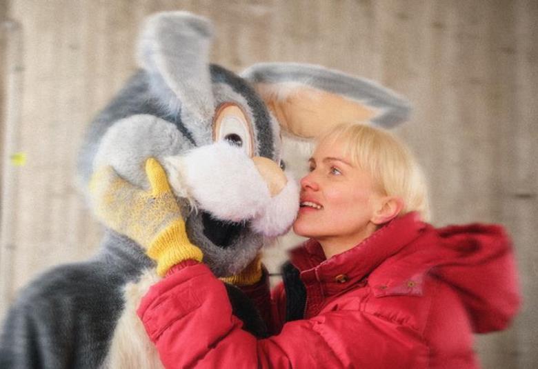 Kuvassa on nuori nainen oikealla punaisessa toppatakissa puolivartalokuvassa. Hän pitää käsiään harmaan vaalean kanin puvussa olevan vähän pidemmän näyttelijän posken päällä ja poski kanin posken päällä. Taustalla on harmaata betoniseinää.