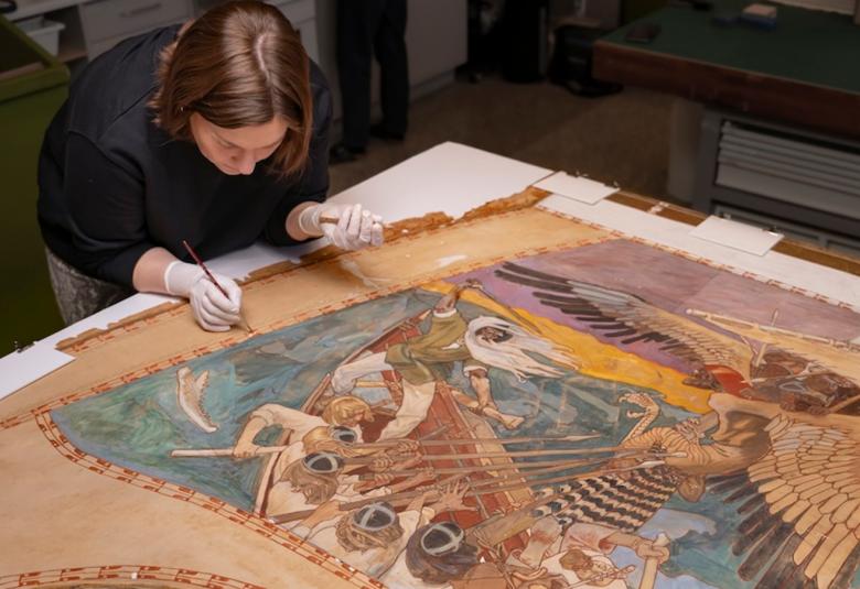 Kuvassa on pöydän ääressä nainen kumartuneena ja valkoisissa hanskoissa oikeassa kädessään sivellin ja konservoi kangasta, jolla on teos.  Siinä on reunoilla vaalean ruskeaa ja turkoosin sekä ruskean sävyinen kuvitus Sammon puolustuksesta.