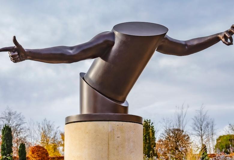 Kuvassa on tumma teräksestä tehty teos, jossa on kaksi lieriötä vinosti päällekkäin etukenossa. Lieriötä lähtee levitetyt kädet. Teos on vaalealla pyöreällä jalustalla. Taustall on sinivalkoista taivasta. 