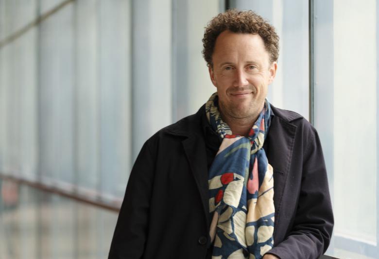 Kuvassa Krist Gruijthuijsen puolivartalokuvassa nojaamassa kaiteeseen harmahtavassa käytävässä, jossa on vaaleata seinää oikealla. Hänellä on musta pusero päällään ja kirjava huivi kaulassaan.