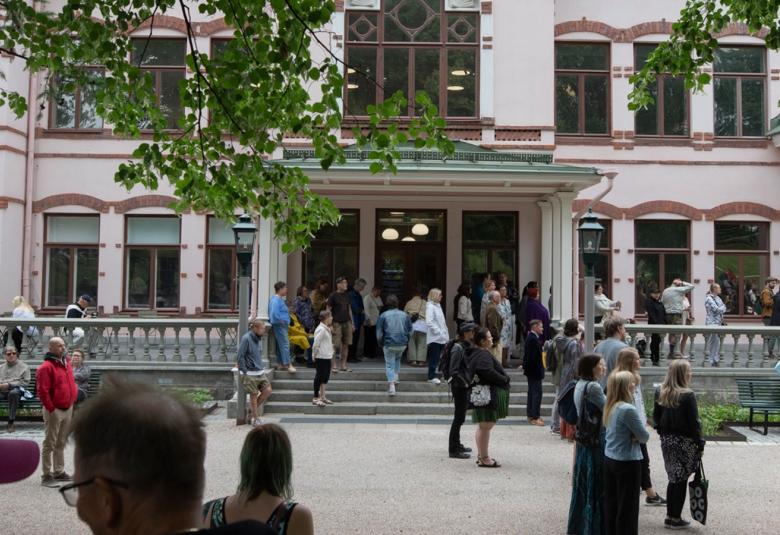 Kuvassa on vaalean roosan värinen Lahden historiallinen museo, jossa on korkeat, kapeat jaetut ikkunaruudut.  Pihassa on ryhmissä paljon ihmisiä seisomassa. Yläpuolella on vihreitä lehtipuun oksia.  