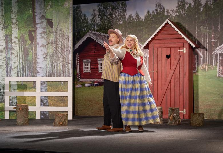 Kuvassa on vasemmalla on koivikkolavastetta ja sen edessä on valkoista aitaa. Oikealla on taustalla punainen talo ja ulkorakennus ja etualalla punainen ulkohuussi. Sen edessä on mies vaalean ruskeassa asussa ja nainen punaisessa liivissä ja ruudullisessa hameessa. 