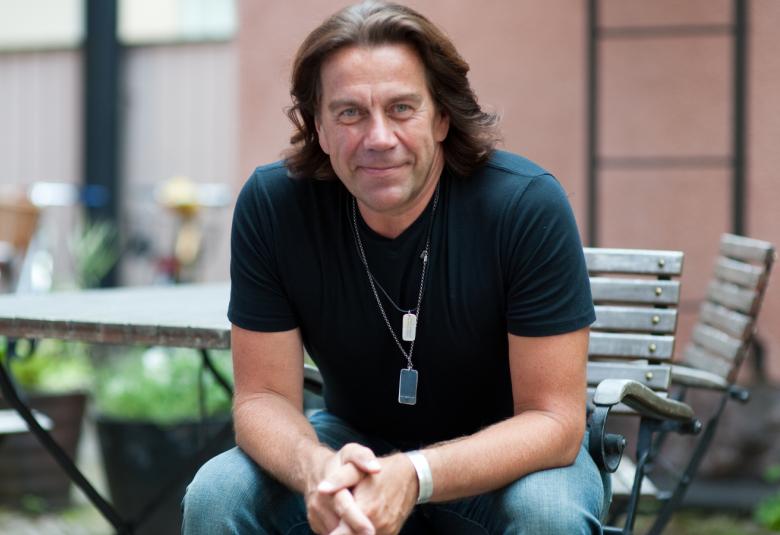 Kuvassa on Mikko Kuustonen istumassa  terassilla, jossa taustalla kesäkalusteita. Kuustonen on kumartuneena eteenpäin kädet ristittyä edessä ja hänellä on sininen t-paita ja farkut päällään.