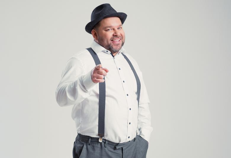 Kuvassa on Mikko Töyssy valkoisessa paidassa, harmaissa henkseleissä ja housuissa musta hattu päässään.  Hän osoittaa oikealla kädellään suoraan eteenpän. Tausta on harmaa.