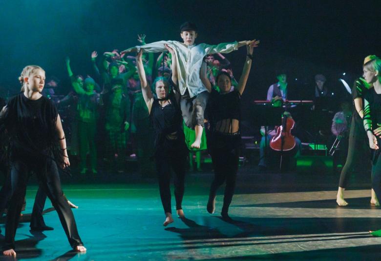 Kuvassa on vasemmalla kolme nuorta tanssijaa mustissa puvuisssa ja oikeassa reunassa toiset tanssijat. Keskellä kaksi tanssijaa, jotka pitävät valkopukuista tanssijaa ylhäällä. 