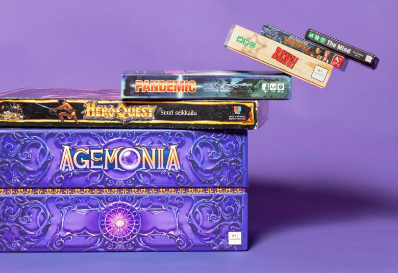 Kuvassa on iso violetti laatikko, jossa lukee kultaisin kirjaimin Agemonia.  Sen päällä on tumma, kapea pelilaatikko ja sen päällä pienempi pelilaatikko, jonka reunasta on ikäänkuin putoamassa kolme pienempää pleilaatikko. Tausta on vaalean violetti.