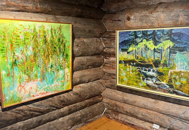 Kuvassa on kaksi taulua kelohirsisellä seinällä kulmittain. Vasemmalla on vaalean vihreää metsää ja oikealla on metsässä virtaavaa vettä sekä tumma taivas.