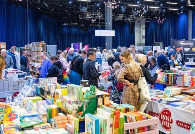 Kuvassa on kirjamessujen yleisöä katsomassa kirjoja täynnä olevien pöytien ääressä kirjoja. Taustalla on sinistä verhoa.