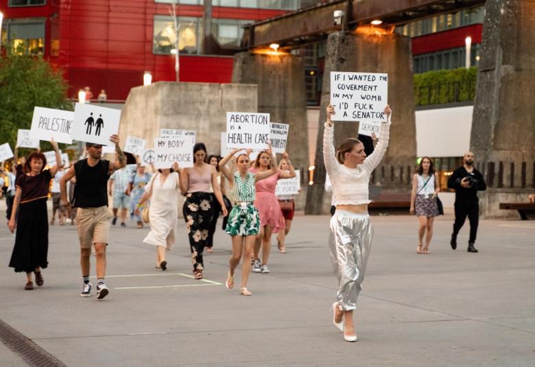 Kuvassa on kesävaatteissa kadulla rakennusten välissä kävelemässä ja heillä on kädessään valkoisia kylttejä, joissa on isoilla kirjaimilla erilaisia tekstejä.  