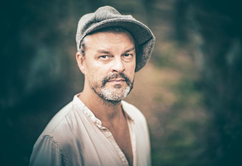 Kuvassa on Tuure Kilpeläinen puolivartalokuvassa. Hänellä on harmaa lippalakki päässään ja vaalea paita päällään.  Tausta on harmaa.