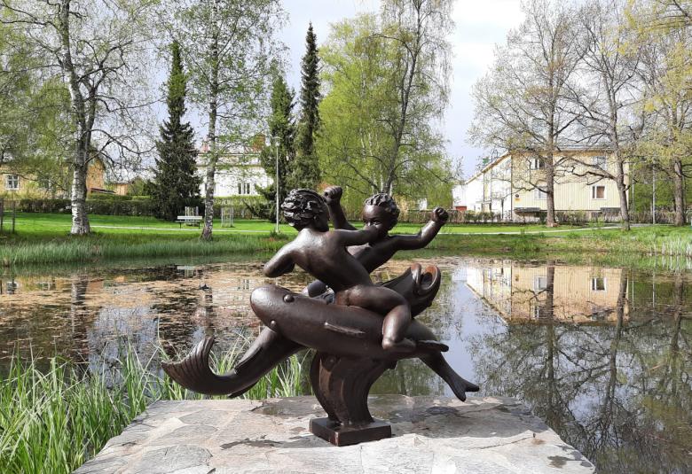 Kuvassa on keskellä lammessa tumma veistos, jossa on kalan päällä kaksi nyrkkeilevää miestä.  Taustalla on näkyy puistoa ja vaalea rakennus ja näkymä heijastuu veteen.
