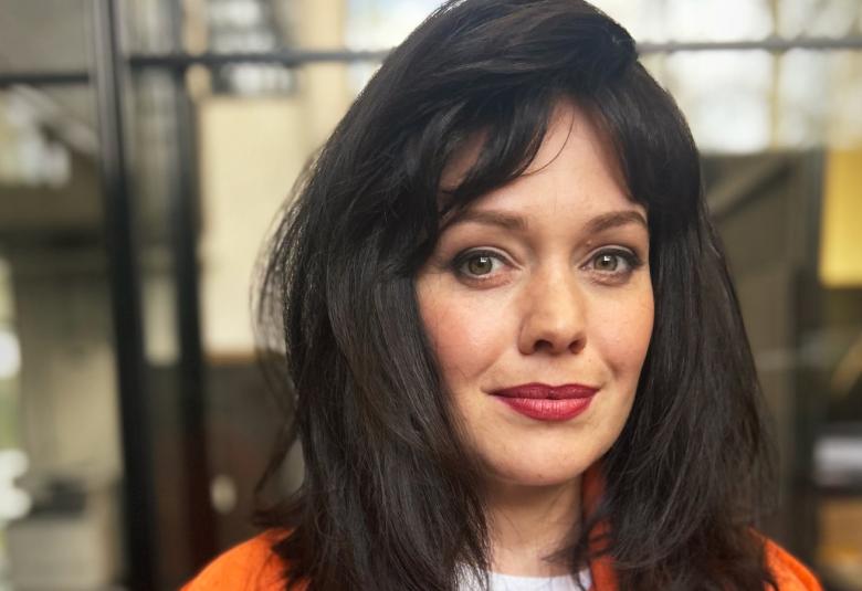 Kuvassa on Oona Airola ja hänellä on tummat pitkät hiukset rooliasussaan. HÄnellä on oranssi pusakka päällään. Taustalla näkyy heijastumia lasi-ikkunasta. 