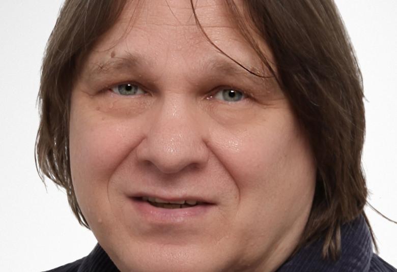 Kuvassa on Jukka Koskelainen kasvokuvassa ja hänellä on tumman ruskeat, puolipitkät hiukset.