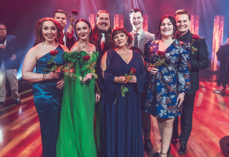 Kuvassa ovat laulajat seisomassa ja jokaisella yksi ruusu kädessään. Edessä ovat naiset ja heistä vasemman puoleisella on turkoosin sininen pitkä puku päällään, viereisellä vihreä puku ja seuraavalla tumman sininen. Oikean puoleisella on polvipituinen sininen perhoskuvioitu mekko.  Takarististä näkyvät mieslaulajien kasvot.  Taustalta näkyy sinistä ja punaista valaistusta.