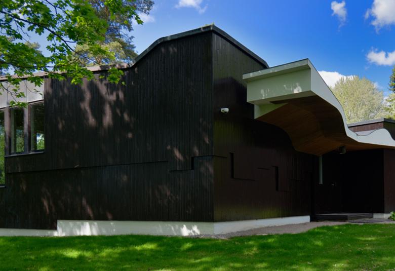 Kuvassa on tumma talo Villa Kokkonen ja siinä on vasemmalla pystyikkuinoita. Katossa on kaarevia muotoja ja seinät ovat rummia. Oikealla on matalampi osa, jossa on pieni ikkuna. Vasemmalla on pihlajan oksia ja taustalta näkyy sinistä taivasta.