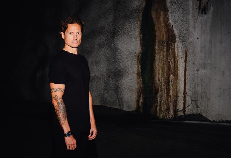 Kuvassa on seisomassa Alexi Tuomarila mustassa, lyhythihaisessa t-paidassa. Taust aon tumma ja siinä harmaata keskellä.