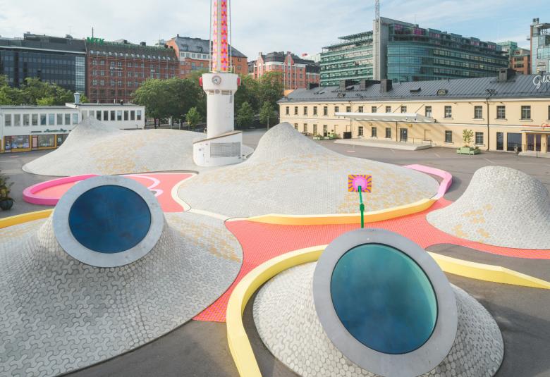 Kuvassa on harmaiden kukkulamaisten kartioiden väliin maalattu roosan väristä maalia ja väleihin sijoitettu keltaiset pötkylät.