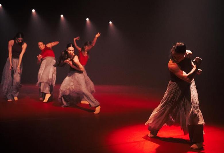 Kuvassa on vasemmalla neljä naistanssijaa harmaissa tyllihameissa ja vasemmalla yksi tanssija käsi oikea käsi kiedottuna olkapäätä vasten ja pää painettuna alaspäin. He tanssivat punaisessa valossa tummalla taustalla.